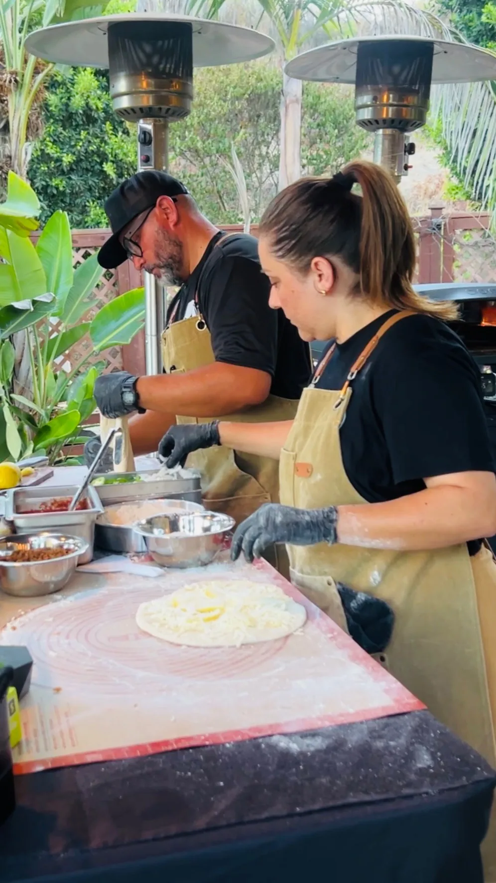 Two people building pizzas side by side at the prep table.